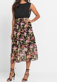 Černé šaty bez rukávů s přiléhavým živůtkem a splývavou sukní s květinovým vzorem v odstínech růžové a zelené, s vyznačeným pasem.