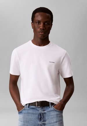 Jeune homme portant un t-shirt blanc à manches courtes Calvin Klein et un jean bleu, les mains dans les poches, sur fond gris.