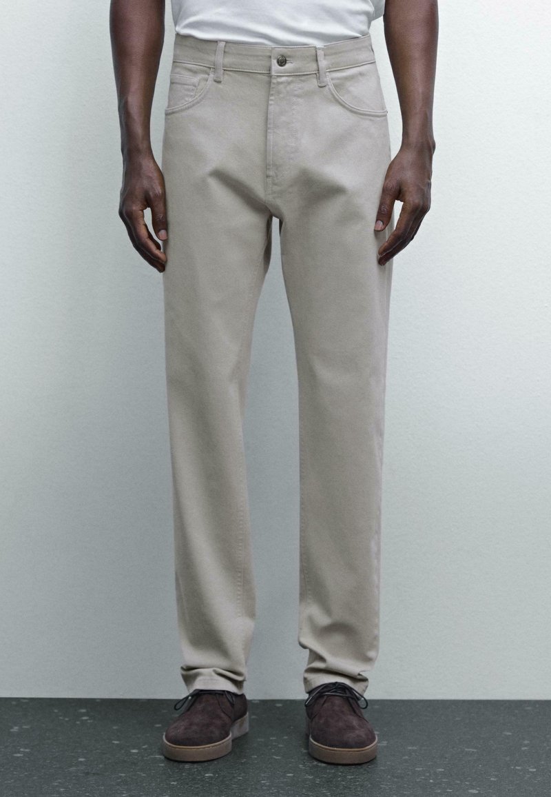 Pantaloni in cotone beige con un design a gamba dritta, dotati di tasche frontali e dettagli di cucitura sottili. Indossati con scarpe in suede marroni.