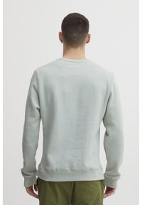 Világosszürke pulóver, kerek nyakkivágással, bordázott mandzsettákkal és aljjal, sima textúrával, hátulról látható egy zöld nadrágot viselő személyen.