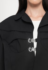 Chaqueta negra con un cuello estructurado, dobles bolsillos en el pecho y cierres de hardware transparente únicos. Tela suave con un diseño minimalista.