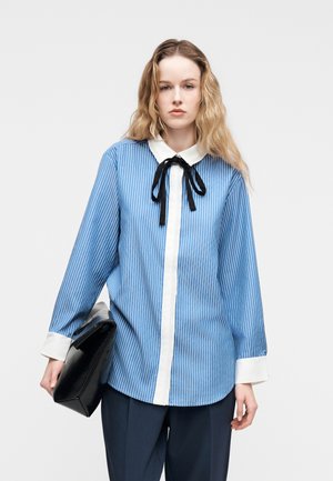 Γυναίκα με μακριά κυματιστά μαλλιά που φοράει μπλε και λευκό πουκάμισο με ριγέ μοτίβο και μαύρη κορδέλα στη γραβάτα, κρατώντας μαύρο τσαντάκι, στέκεται μπροστά σε απλό φόντο.