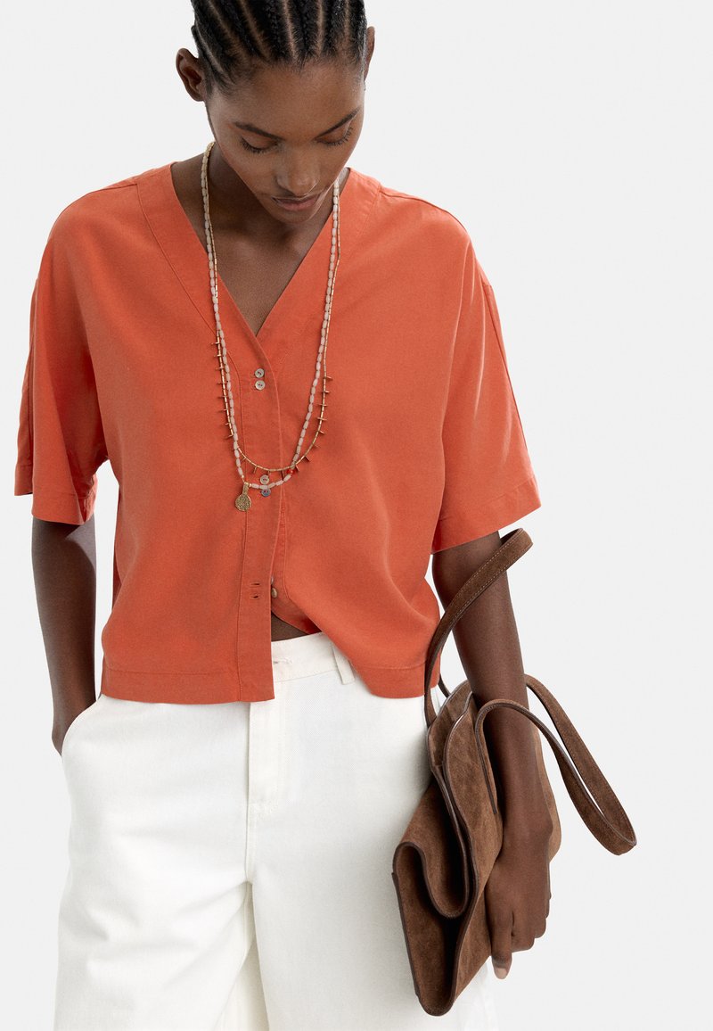 Koszulka z krótkim rękawem w kolorze palonej pomarańczy, z dekoltem w kształcie litery V, z dwoma guzikami i krótkim krojem, zestawiona z białymi spodniami i brązową torbą z zamszu.