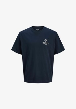 Băiat purtând șapcă roșie, tricou bleumarin, geacă din denim legată în jurul umerilor, blugi largi albaștri și pantofi bej, stând în fața unui fundal simplu.
