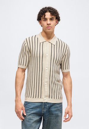 Ung mand med krøllet hår iført en beige kortærmet skjorte med lodrette sorte striber og blå jeans foran en ensfarvet baggrund.