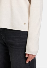 Jasno beżowy dziergany sweter o teksturowanym wykończeniu; posiada złoty akcent na dolnej krawędzi; zestawiony z czarnymi dżinsami.