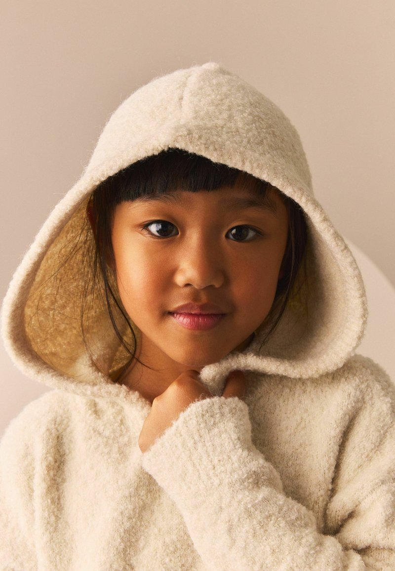 Haut en sweatshirt blanc texturé avec une grande capuche, en tissu confortable, et des poignets côtelés, porté par un enfant. Des tons naturels mettent en valeur les traits du visage et la chaleur.