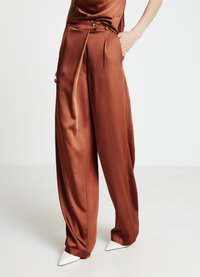 Satin-Hose mit weitem Bein in Rostfarbe, mit hohem Bund und goldfarbenen Beschlägen. Kombiniert mit spitzen weißen Schuhen.