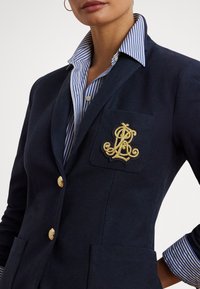 Blazer azul-marinho com botões dourados e um emblema no bolso. Por baixo, é visível a gola de uma camisa às riscas azuis e brancas.