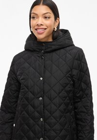 Pufasta črna jakna s kapuco, ki ima zaklopne gumbe in stranske žepe, izdelana iz gladkega blaga z diamantnim vzorcem.