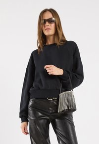 Schwarzer übergroßer Sweatshirt aus weichem Stoff, kombiniert mit glänzenden schwarzen Lederhosen. Die Accessoires umfassen eine fransige metallische Mini-Tasche und getönte Sonnenbrillen.