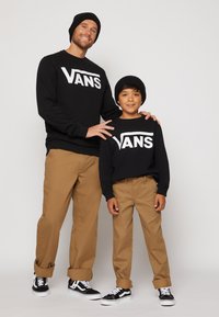 Zwarte sweaters met het "VANS" logo, tan broek en zwarte beanies. Beiden dragen zwart-witte sneakers met vetersluiting.