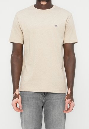 Homme atteint de vitiligo portant un t-shirt beige à manches courtes et un jean gris, debout devant un fond blanc uni.