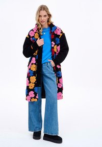 Farebný kvetinový vzorovaný čierny kabát cez modrú košeľu, doplnený o široké džínové nohavice a čierne platformové topánky. Mäkká textura.