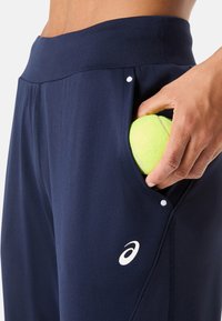 Navy sportbroeken met een zachte textuur, voorzien van een grote zijklep met een felgele tennisbal erin en een wit logo op de bovenbeen.