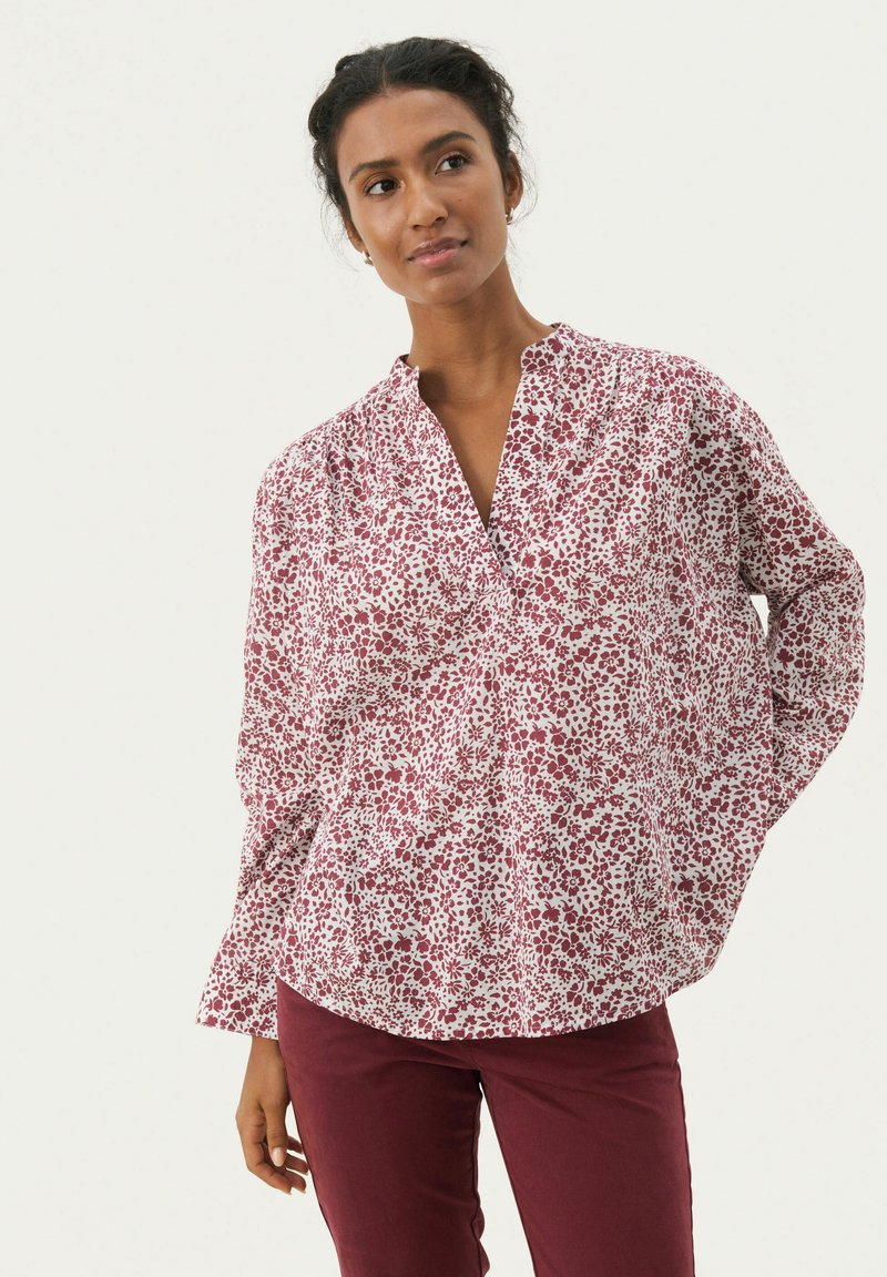 Long-sleeve blouse in white with a red floral pattern. Features a notch collar and loose fit, made from lightweight fabric. Paired with burgundy pants.