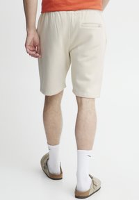 Pantalones cortos de algodón beige con cintura elástica y un bolsillo trasero, que presentan una textura suave y un corte relajado, combinados con zapatillas grises.