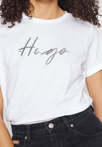 Vit bomullströja med uppvikta ärmar som har en silverfärgad metallisk handskriven "Hugo"-logotyp. Matcher med svarta denimjeans.