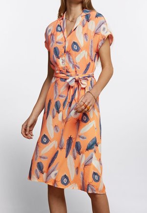 Femme portant une robe pêche mi-longue avec un imprimé de plumes bleu et blanc, manches courtes, devant boutonné et ceinture nouée à la taille.