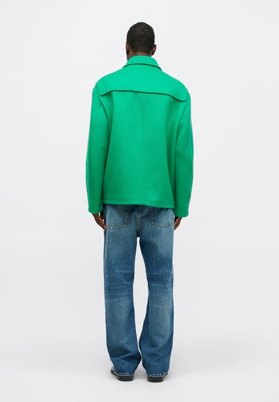 Chaqueta verde de gran tamaño con un amplio cuello, hecha de un material suave y texturizado. Combinada con unos vaqueros de denim azul de corte holgado.