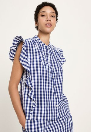 Woman wearing blue and white checkered sleeveless button-up blouse with ruffle trim and matching skirt, standing with one hand in pocket.