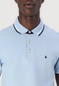 Lyseblå polo t-shirt med krave, tre knaplukninger og marineblå stribede detaljer ved kraven. Har et lille logo på venstre side.