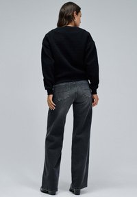 Černý svetr s texturou a širokými rukávy, v kombinaci s tmavě šedými džíny s vysokým pasem a zadními kapsami, doplněný černými kotníkovými botami.