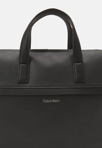 Borsa tote in pelle nera con superficie testurizzata, caratterizzata da due robuste maniglie e un design minimalista. Logo "Calvin Klein" in argento.