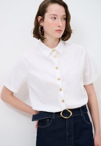 Camicetta bianca a maniche corte con colletto classico e bottoni dorati; indossata con una gonna di jeans scura caratterizzata da una fibbia d'oro testurizzata.