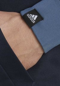 Blue cuff with visible stitching and a black label featuring the Adidas logo, paired with a dark fabric underneath. Skin tone is visible in the hand.