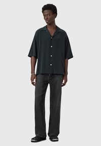 Chemise noire à manches courtes à boutons, avec une coupe décontractée, associée à un jean en denim sombre et large, ainsi qu'à des chaussures noires avec des accents blancs.
