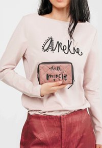 Cartera acolchada rosa con cremallera y detalles negros, que presenta texto decorativo. Se lleva con una camiseta de manga larga rosa claro que tiene un logotipo.