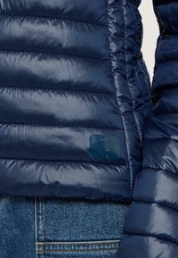 La giacca puffer blu navy presenta un design trapuntato, una texture liscia e una patch con il logo. Indossata sopra dei jeans in denim blu, evidenziando il look a strati.