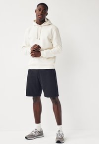 Haut à capuche de texture crème avec cordons de serrage, associé à un short noir. Chaussures de sport grises et blanches avec des chaussettes blanches à côtes. Design minimaliste.