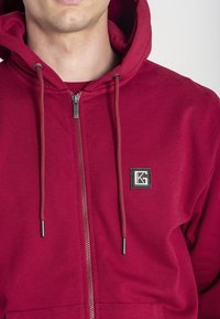 Bordová mikina na zips s kapucňou na stiahnutie, mäkká tkanina, predné vrecká a malá štvorcová nášivka na hrudi.