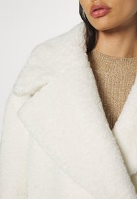 Femme portant un manteau en laine blanche texturée sur un pull en maille marron clair, vue des épaules aux lèvres.