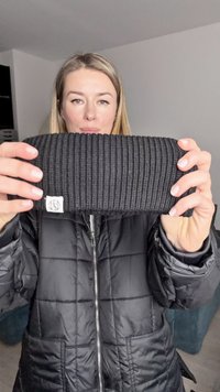 Bandeau en tricot noir côtelé avec une petite étiquette blanche, tenu par une personne portant une doudoune noire. Le design est simple et épais.