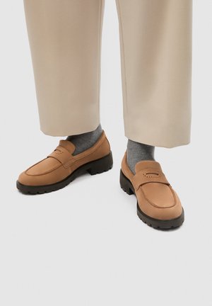 Voeten met beige broek, grijze sokken en lichtbruine suède loafers met zwarte grove zolen op een witte achtergrond.