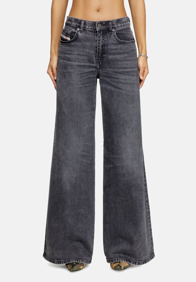 Jean en denim noir délavé à jambe large porté par une personne debout, montrant les poches avant, les passants de ceinture et des chaussures camouflage pointues.