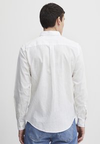 Camisa blanca de manga larga hecha de lino ligero y texturizado, con dobladillo curvado y puños abotonados; presenta acentos de botones amarillos.