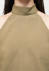 Γυναίκα που φοράει μπεζ τοπ χωρίς μανίκια με ψηλό λαιμό και ανοίγματα στους ώμους, αποκαλύπτοντας το λαιμό, τις κλείδες και τα μαλλιά της.