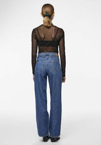 Top lungo in rete nera a maniche lunghe indossato sopra un cinturino nero aderente, abbinato a jeans a vita alta blu con gambe larghe e tasche posteriori.