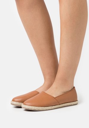 Legs wearing tan slip-on espadrille flats with woven rope soles, standing on a white background.