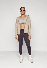 Beige hettejakke med glidelås, grå sports-BH med mønster, svarte tights og kremfargede joggesko. Modellen har på seg store solbriller og står i en nøytral posisjon.