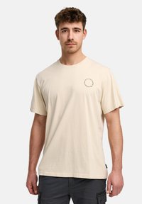 Camiseta de algodón de color beige claro con mangas cortas y cuello redondo. Presenta un pequeño diseño impreso circular en el lado izquierdo del pecho.
