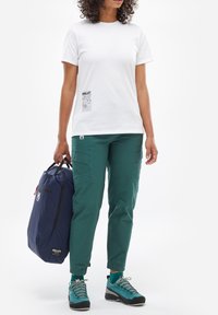 T-shirt en coton blanc avec un imprimé graphique, associé à un pantalon cargo vert et des chaussures de randonnée teal. Un sac navy est tenu dans une main.