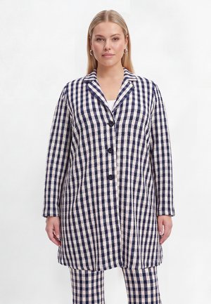 Vrouw draagt een lange marineblauwe en witte geruite jas met zwarte knopen en bijpassende geruite broek, staand tegen een effen witte achtergrond.