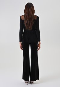 Tuta nera con maniche lunghe e pannello posteriore trasparente, caratterizzata da un corpetto aderente e pantaloni svasati, con texture liscia e design semplice.