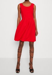 Robes sans manches rouges avec un corsage ajusté et une jupe évasée. Tissu lisse, détails de plis et encolure ronde. Sandales à talons noirs avec des lanières.