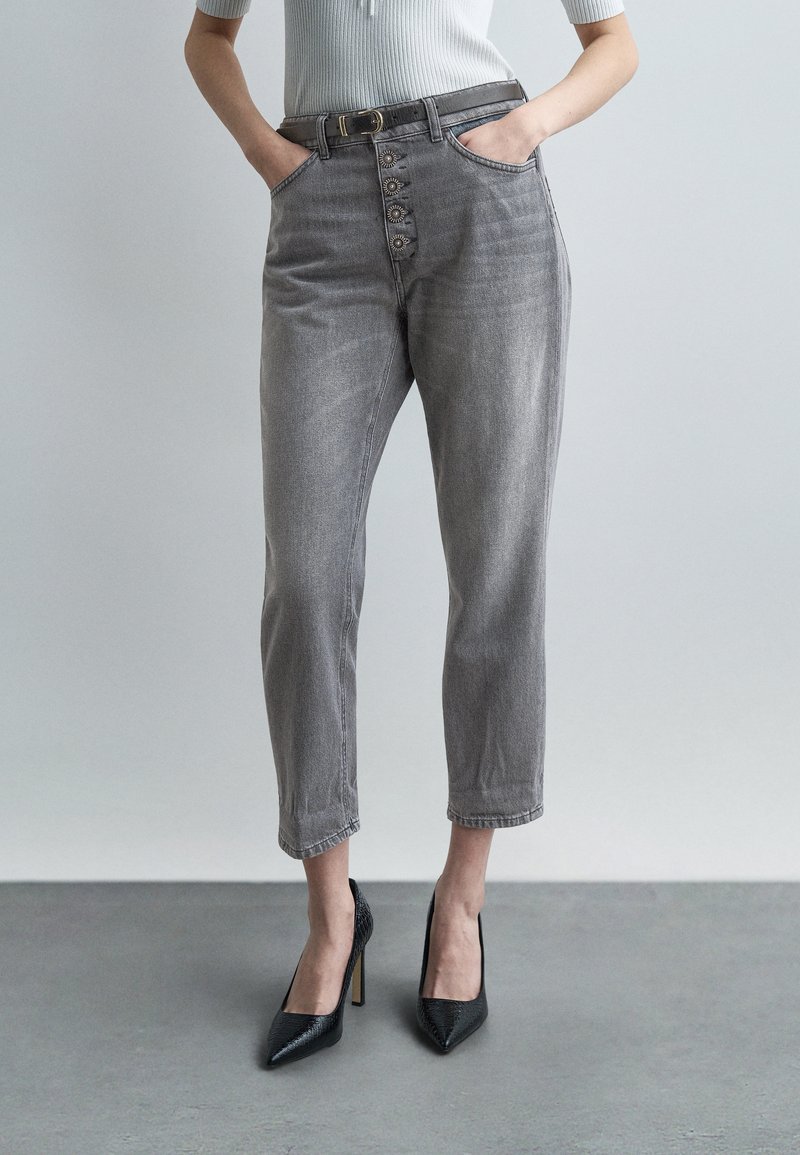 Γυναίκα που φοράει γκρι τζιν ψηλόμεσο με διακοσμητικά κουμπιά, μαύρη ζώνη, ανοιχτό γκρι τοπ και μαύρα ψηλοτάκουνα παπούτσια, στέκεται σε εσωτερικό χώρο.